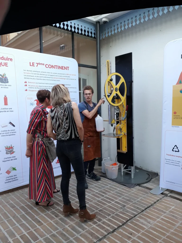 Deux collaboratrices écoutent l’animateur qui présente l’extrudeuse pour créer un objet en plastique recyclé, sur une activité team building green.