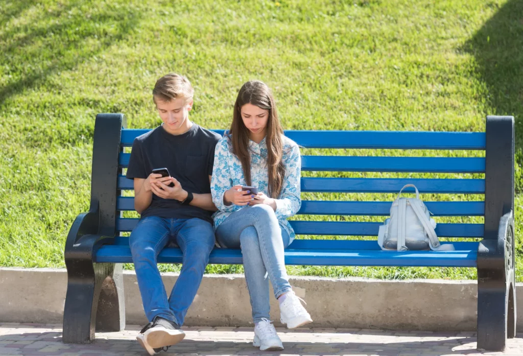 2 jeunes consultent leurs téléphones assis sur un banc public.